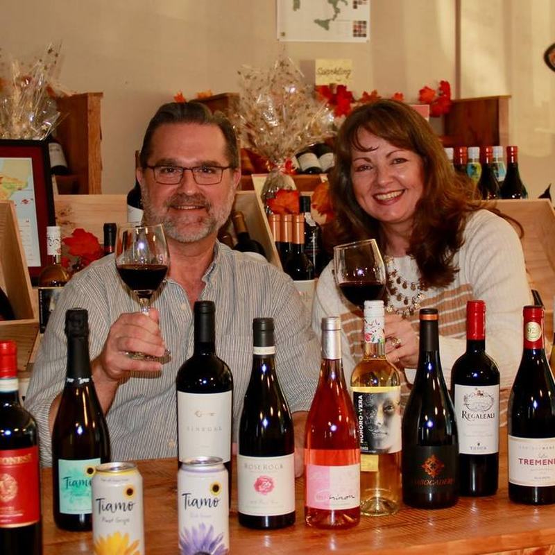 Owners Kirk and Candy raising glasses of red wine behind a counter lined with bottles