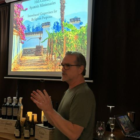 Kirk presenting a wine class in front of a projected slide, with bottles lined up on the table in front of him