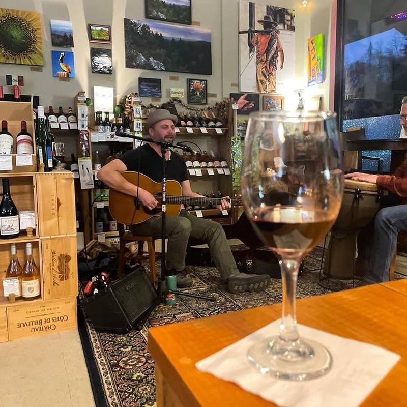 A guitarist and a percussionist playing live music inside Bryson City Wine Market at night, seen past a glass of wine on the tasting bar